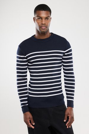 Mann trägt einen marineblauen Pullover mit weißen horizontalen Streifen und schwarze Hosen, steht vor einem einfarbigen hellen Hintergrund.