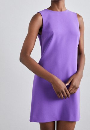 Personne portant une robe violette sans manches, longueur genou, debout les mains doucement jointes devant un fond clair uni.