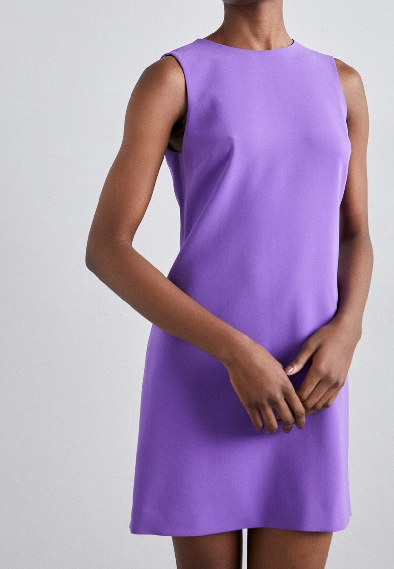 Personne portant une robe violette sans manches, longueur genou, debout les mains doucement jointes devant un fond clair uni.