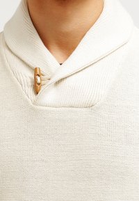 Beżowy sweter z dzianiny z kołnierzem typu szal i zapięciem na drewniany toggle. Teksturowany materiał z poziomymi detalami szycia.