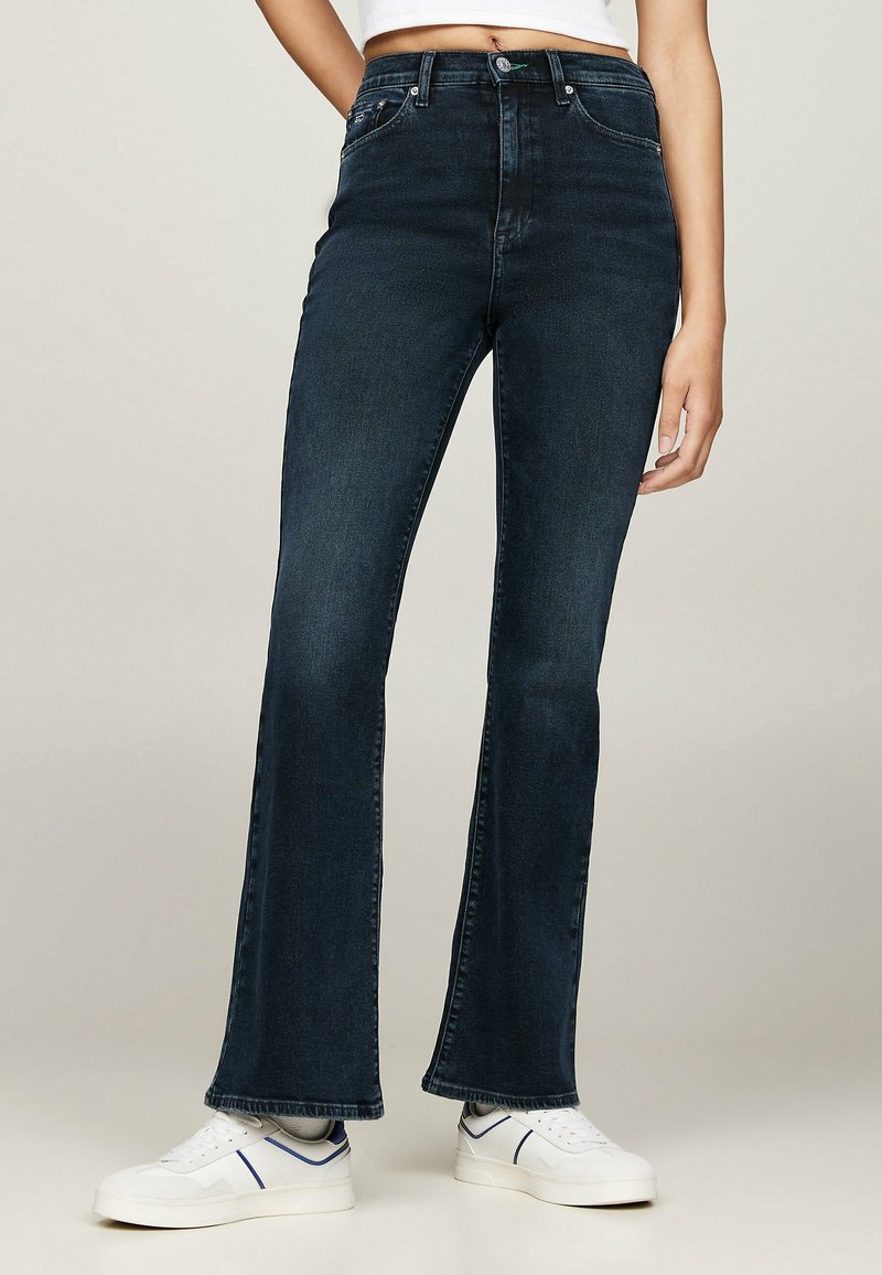 Kobieta ubrana w ciemnoniebieskie jeansy z wysokim stanem i rozszerzanymi nogawkami oraz białe trampki z niebieskimi akcentami, stojąca na neutralnym tle.