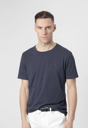 Joven con cabello corto y oscuro, vistiendo una camiseta lisa azul marino y pantalones blancos, de pie frente a un fondo gris claro.