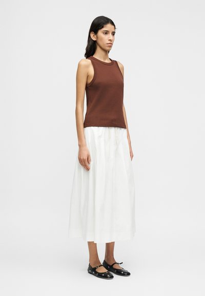 Καφέ, εφαρμοστό τοπ από βαμβάκι· συνδυασμένο με λευκή, αέρινη, πλισέ φούστα. Μαύρα, επίπεδα παπούτσια με διακοσμητικά μεταλλικά στοιχεία.