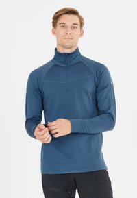 Maglietta a maniche lunghe con zip nel colletto, di colore blu scuro, realizzata in tessuto leggero e liscio. Presenta cuciture a contrasto e un design aderente.