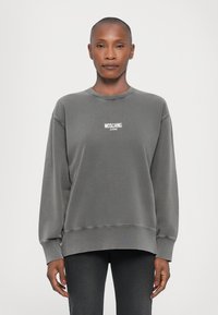 Sudadera gris con cuello redondo, hombros caídos y puños de canalé. Cuenta con el logo blanco "MOSCHINO JEANS" en el pecho. Textura de tela suave.