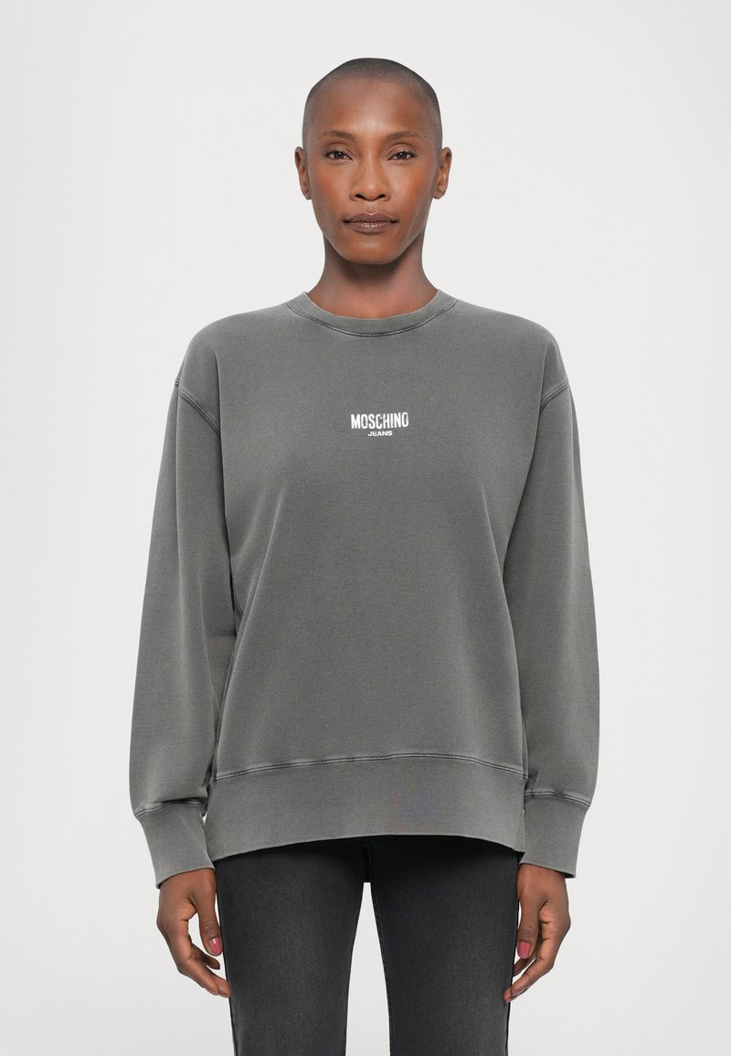 Sudadera gris con cuello redondo, hombros caídos y puños de canalé. Cuenta con el logo blanco "MOSCHINO JEANS" en el pecho. Textura de tela suave.