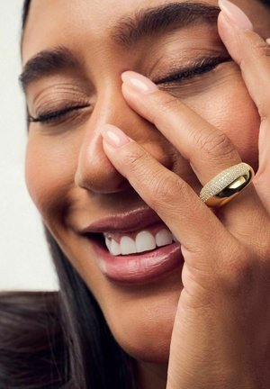 Guldring med en tekstureret overflade og glatte kanter, med et bånddesign. Nærbillede af en hånd, der dækker en del af et smilende ansigt.