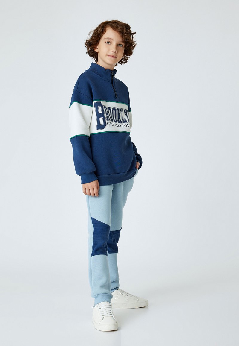 Marinblå zip-hoodie med texten "BROOKLYN", vita och gröna ränder. Ljusblå träningsoverallsbyxor med marinblå detaljer och strukturerade paneler. Vita sneakers.