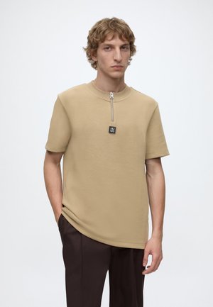 Junger Mann mit lockigem Haar, der ein beige kurzärmeliges Hemd mit Frontreißverschluss und dunkelbraune Hose trägt, steht mit einer Hand in der Tasche.