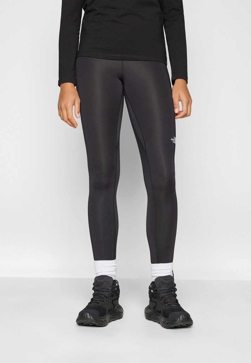 Fekete sportleggings sima textúrával, testhez álló szabással és fehér logóval a boka közelében. Fekete sportcipővel párosítva.