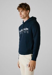 Marineblauwe hoodie met een logo op de voorkant, gemaakt van zachte stof. Het ontwerp bevat een kangoeroe zak en een verstelbare trekkoordkap.