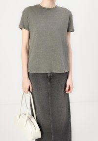 Zielono-oliwkowy t-shirt z krótkim rękawem o przecieranym wyglądzie, zestawiony z ciemnoszarym dżinsowym spódnicą. Trzyma białą torebkę o strukturalnym kształcie.