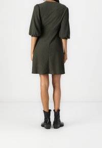 Vestido corto verde oscuro con mangas abullonadas, tejido texturizado, cuerpo ajustado y falda acampanada, combinado con botas negras hasta el tobillo.