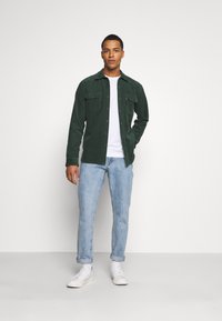 Camisa de cortiça verde com fecho de botão, dois bolsos no peito, camiseta branca de cuello redondo por baixo, jeans azul claro e ténis brancos.