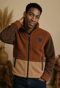 Bruine en tan fleece jas met een ritssluiting aan de voorkant, hoge kraag en twee zijzakken. Bevat contrasterende kleurvlakken en zwarte accenten.