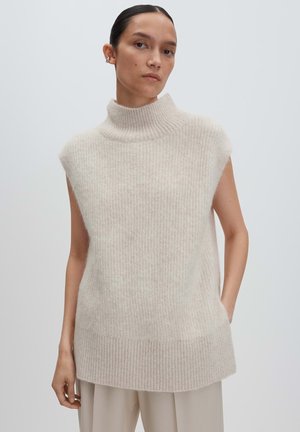 Femme aux cheveux foncés portant un débardeur en col roulé beige côtelé sans manches et un pantalon de couleur claire, se tenant devant un fond uni.