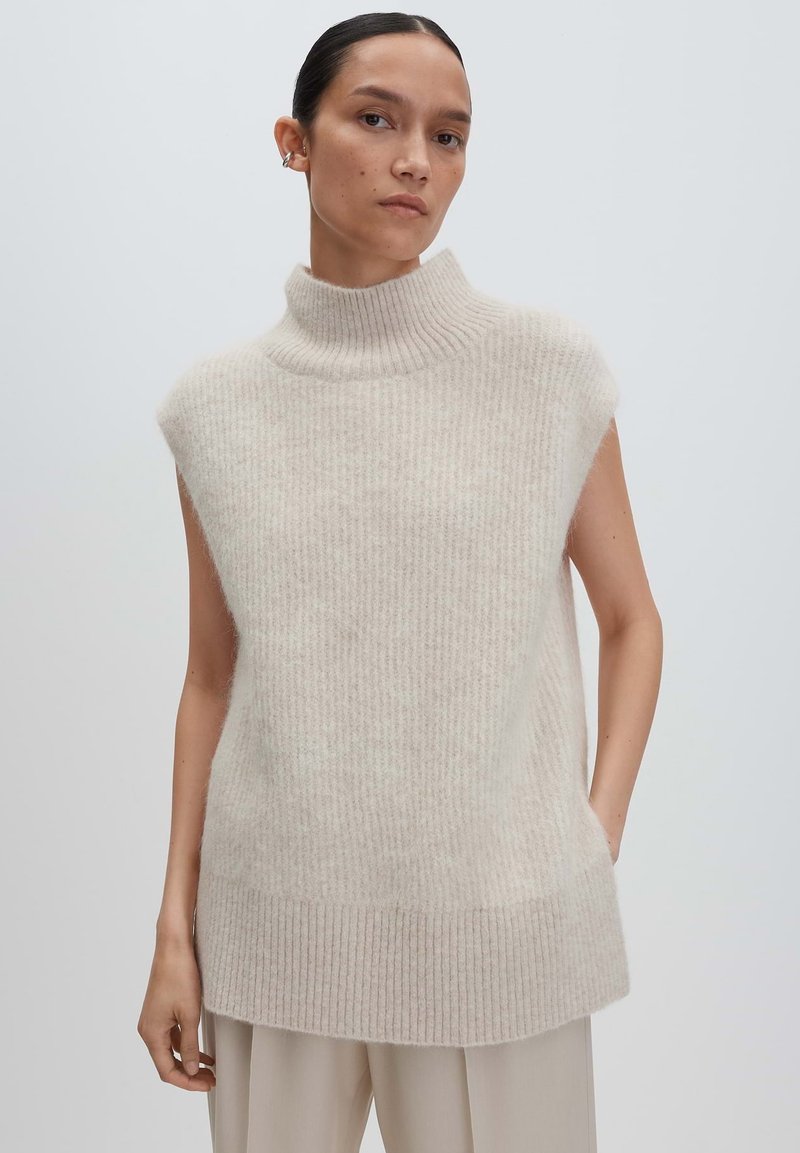 Femme aux cheveux foncés portant un débardeur en col roulé beige côtelé sans manches et un pantalon de couleur claire, se tenant devant un fond uni.