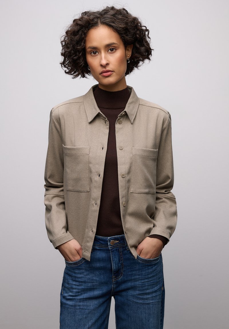 Beiges Hemd zum Knöpfen mit zwei Brusttaschen, getragen über einem braunen Rollkragenpullover, kombiniert mit blauen Jeans. Glatte Textur, klassisches Kragen-Design.