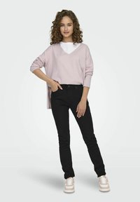 Un pull v-neck surdimensionné rose pâle superposé sur une chemise blanche, associé à un jean skinny noir et des baskets roses, se tenant devant un fond uni.