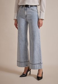 Wide-leg light blue denim jeans with a raw hem, detailed stitching, and a high waist, paired with black high-heeled shoes.