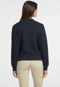 Sweatshirt in cotone blu scuro con scollo rotondo, polsini e orlo a coste. Indossato da un modello che porta pantaloni beige attillati.