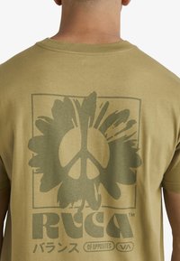 Camiseta de algodón verde oliva con un gran gráfico de un símbolo de paz dentro de un diseño floral, contorneado con un borde cuadrado.