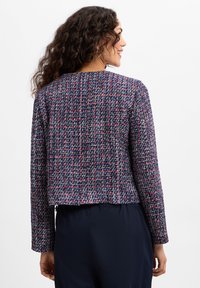 Marineblauwe tweedjas met een structuurpatroon, met rode en witte accenten. Gekorte stijl met lange mouwen en ronde halslijn.