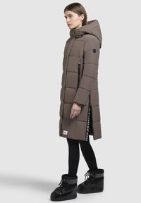 Dugi, podstavljeni parka u zemljanoj smeđoj boji s kapuljačom, vertikalnim crnim patentnim zatvaračima i logo naglascima. Crne tajice i sjajne crne čizme upotpunjuju izgled.