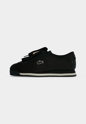 Czarny sneaker Lacoste o niskim profilu, podeszwa z białym brzegiem, wyszywane logo krokodyla po boku oraz czarne sznurówki ze złotymi przelotkami.