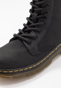 Black high-top boot featuring a textured fabric upper, rugged rubber sole with yellow accents, and black laces through metal eyelets.