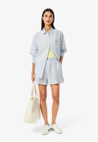 Camisa de rayas azul claro con manga larga, combinada con pantalones cortos a juego. El modelo sostiene una bolsa de tela color crema y lleva zapatillas blancas.