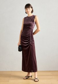 Μπορντό βελούδινο midi φόρεμα με εφαρμοστή κορυφή, λεπτομέρεια ραφής στο πλάι και ίσια σιλουέτα, συνδυασμένο με μαύρα αξεσουάρ και σανδάλια ανοιχτού σταδίου.