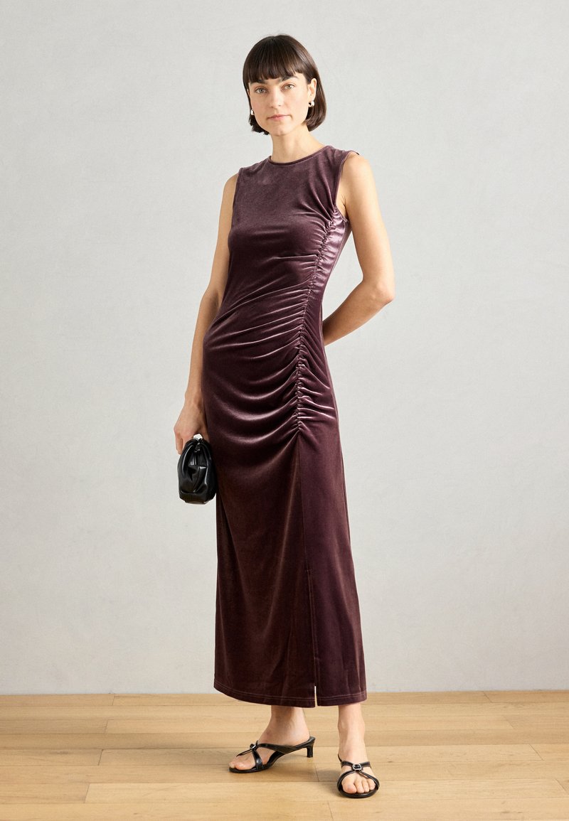 Μπορντό βελούδινο midi φόρεμα με εφαρμοστή κορυφή, λεπτομέρεια ραφής στο πλάι και ίσια σιλουέτα, συνδυασμένο με μαύρα αξεσουάρ και σανδάλια ανοιχτού σταδίου.