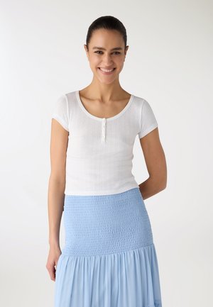 Donna sorridente con capelli scuri raccolti, che indossa una maglia bianca a maniche corte e una gonna azzurro chiaro con trama, su uno sfondo semplice.