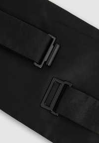 Musta kangaskassi, jossa on kaksi säädettävää hihnaa ja mustaa metalliosaa. Ominaisuuksina on rakenne, jossa on teksturoitu pinta ja litteä, suorakulmainen muoto.
