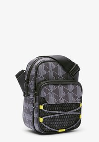 Pequeño bolso bandolera negro con estampado geométrico blanco, que cuenta con dos compartimentos con cremallera, panel texturizado negro y correas de acento amarillas.