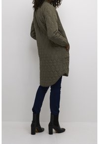 Pikowana zielona kurtka z długimi rękawami, okrągłym dekoltem, bocznymi kieszeniami i asymetrycznym dołem, zestawiona z ciemnymi dżinsami i czarnymi botkami na obcasie.