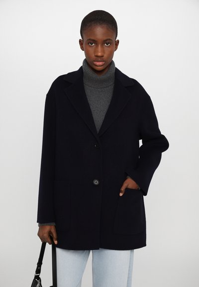 Manteau en laine navy avec revers en pointe, fermeture à un bouton, deux poches avant et coupe décontractée, porté par-dessus un pull col roulé gris.