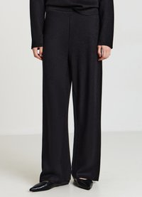 Schwarze weite Hosen aus einem weichen, strukturierten Stoff. Sie haben eine hohe Taille und reichen bis zum Boden, kombiniert mit spitzen schwarzen Ballerinas.