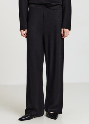 Schwarze weite Hosen aus einem weichen, strukturierten Stoff. Sie haben eine hohe Taille und reichen bis zum Boden, kombiniert mit spitzen schwarzen Ballerinas.