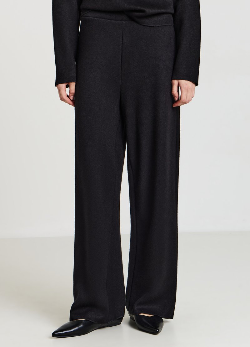 Schwarze weite Hosen aus einem weichen, strukturierten Stoff. Sie haben eine hohe Taille und reichen bis zum Boden, kombiniert mit spitzen schwarzen Ballerinas.