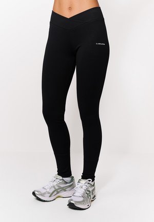 Kobieta w czarnych legginsach z wysokim stanem i paskiem w kształcie litery V oraz białe buty do biegania w zielone wzory, stojąca na białym tle.