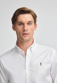 Camicia bianca con bottoni in cotone, con un piccolo logo blu di un golfista sul lato sinistro del petto, dotata di un colletto classico e dettagli con bottoni.