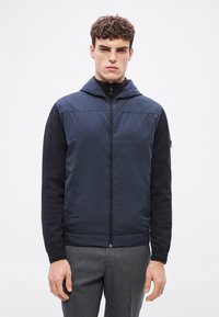 Tumši zila kapuce jaka ar polsterētu priekšpusi un garām melnām piedurknēm; aprīkota ar divām priekšējām kabatām un rāvējslēdzēja aizdari; gluda tekstūra.