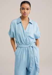Lys blå, kortærmet omslags jumpsuit med krave, bundet talje og sidelommer. Fremstillet af blød, glat stof med en afslappet pasform.