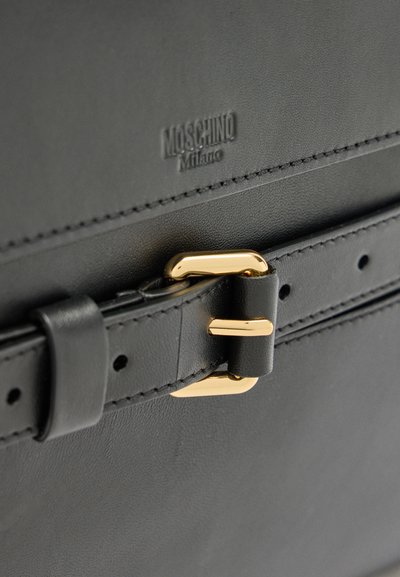 Nærbillede af sort lædertaske med guldlås og præget "Moschino Milano"-logo over detaljer med syet rem.