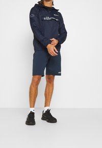 Marinblå vindjacka med framficka och logotyp, kombinerad med mörkblå shorts. Svarta sneakers och vita sportstrumpor fulländar looken.