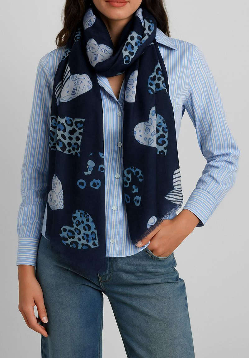 Donna con una sciarpa blu navale con motivi a cuori e animali bianchi e blu sopra una camicia a righe azzurre e jeans blu.