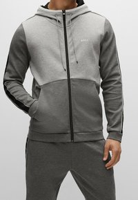 Uomo che indossa una felpa grigia con zip bicolore e pantaloni della tuta abbinati, con strisce nere con logo lungo le maniche e le gambe.