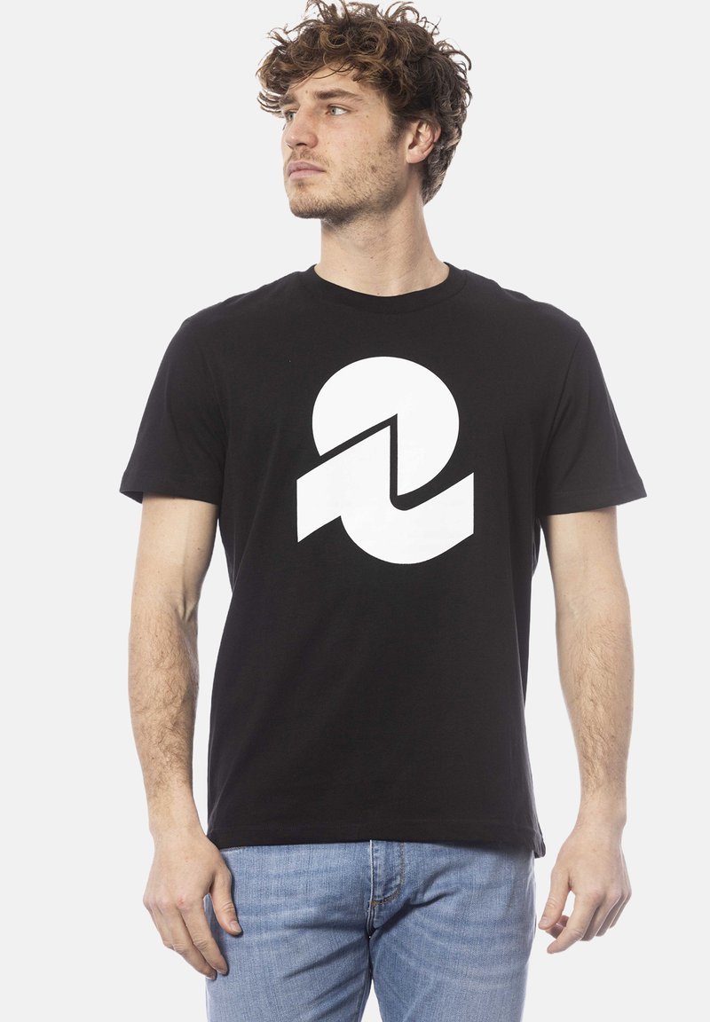 Zwarte katoenen T-shirt met een grote witte grafiek van in elkaar grijpende vormen. Korte mouwen en een standaard ronde halsontwerp. Blauwe jeans zichtbaar.